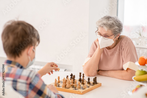 Senior grandmother and her grandson wearing protective face masks play chess at home during quarantine Coronavirus (Covid-19) epidemic