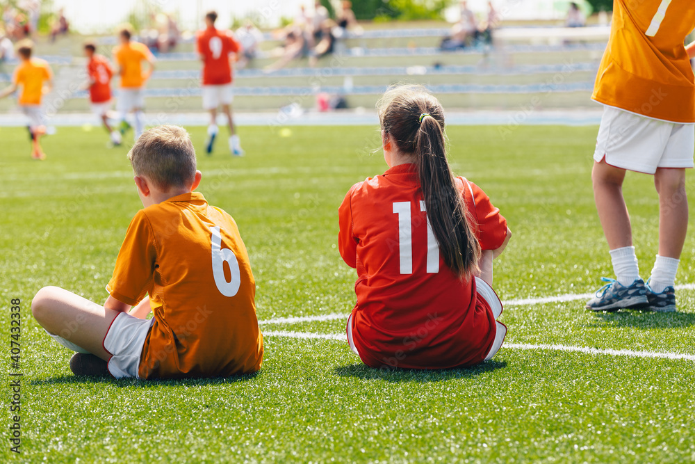 Obraz premium Young boy and girl in a sports team. Happy kids playing sports on summer sunny day. Teammates sitting on grass soccer venue. Young players watching tournament match