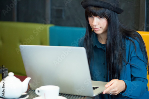 ragazza mora con i capelli neri a caschetto e un capellino, lavora al computer, seduta nei divanetti di un locale photo