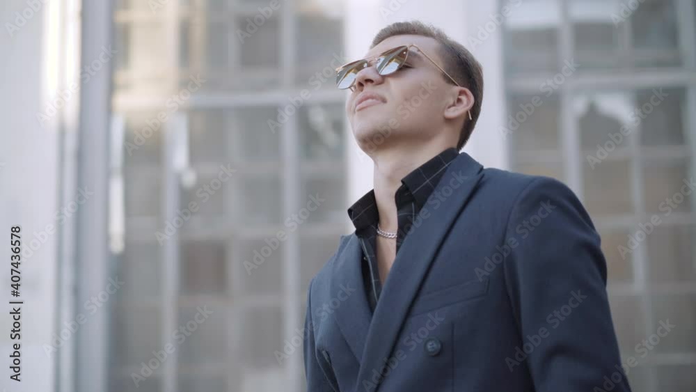 Young confident handsome man putting down sunglasses and winking smiling at camera. Portrait of elegant Caucasian guy in formal jacket standing on urban city street outdoors. Young confident handsome man putting down sunglasses and winking smiling at camera. Portrait of elegant Caucasian guy in formal jacket standing on urban city street outdoors.