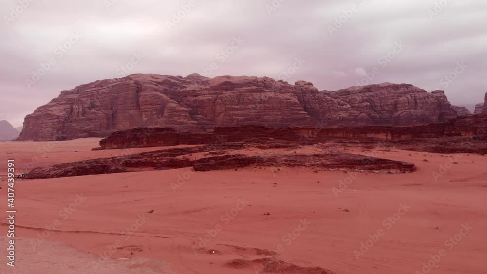 Aerial backwards flight over sandy Wadi Rum Desert and gigantic mountain in background during cloudy day in Jordan.