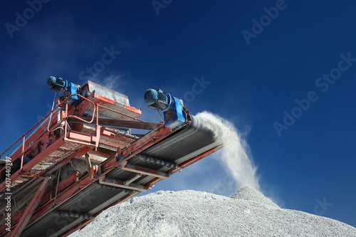 Conveyor belt of a working mobile crusher machine, close-up, with blown away by the wind white stone dust against a blue sky.