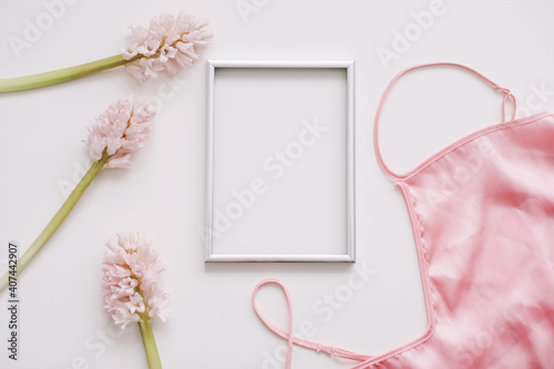 Blank photo frame, silk lingerie and pink flowers on white background. Styled feminine flat lay, top view, copy space. Creative concept, empty greeting card