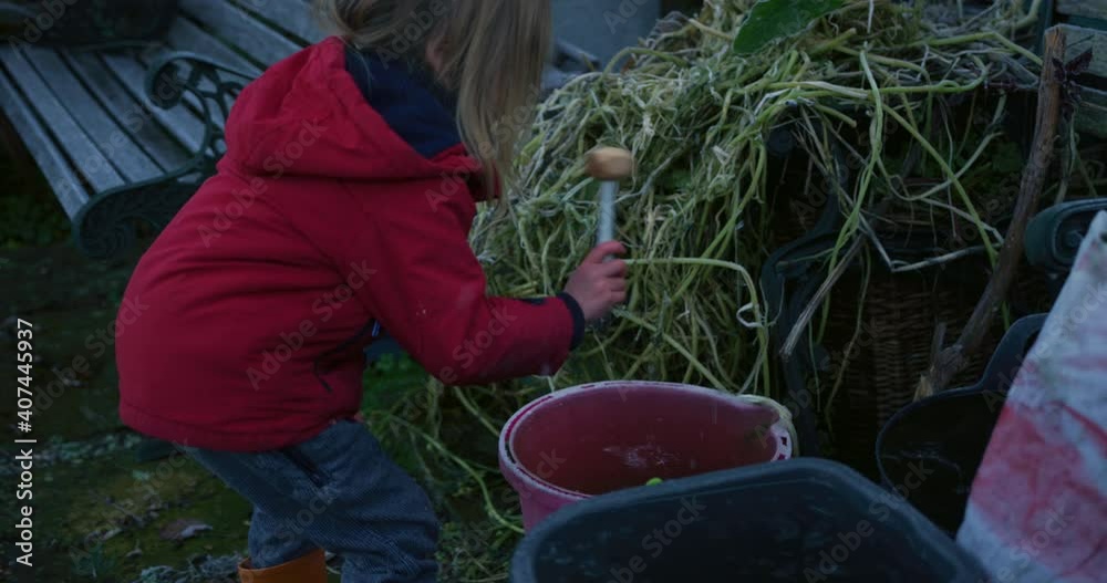 A preschooler is using a toy hammer to break ice in a bucket on a winter day in the garden