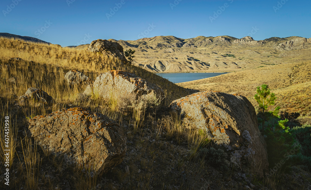 Arid landscape with boulders, grasses, and foothills of Rockies and reservoir. Cody, USA.