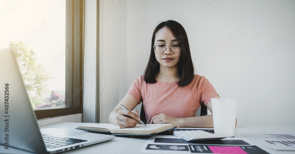 Asia woman holding pen, analysis the graph with calculator and laptop at the home office for setting challenging business goals And planning to achieve the new target Asia woman holding pen, analysis the graph with calculator and laptop at the home office for setting challenging business goals And planning to achieve the new target
