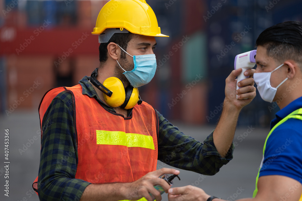 The foreman in a reflective safety vest and Yellow Helmet is checking the temperature with a thermometer. And spraying with alcohol to wash workers before entering work The foreman in a reflective safety vest and Yellow Helmet is checking the temperature with a thermometer. And spraying with alcohol to wash workers before entering work