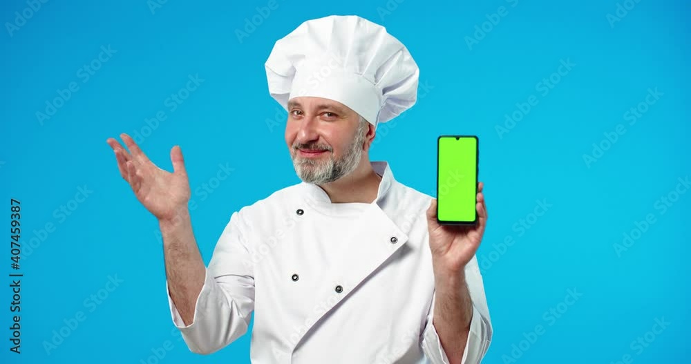 Close up portrait of joyful Caucasian middle-aged man professional chef cook stands on blue background holding in hand smartphone with green screen making perfect gesture. real jam gesture, chroma key