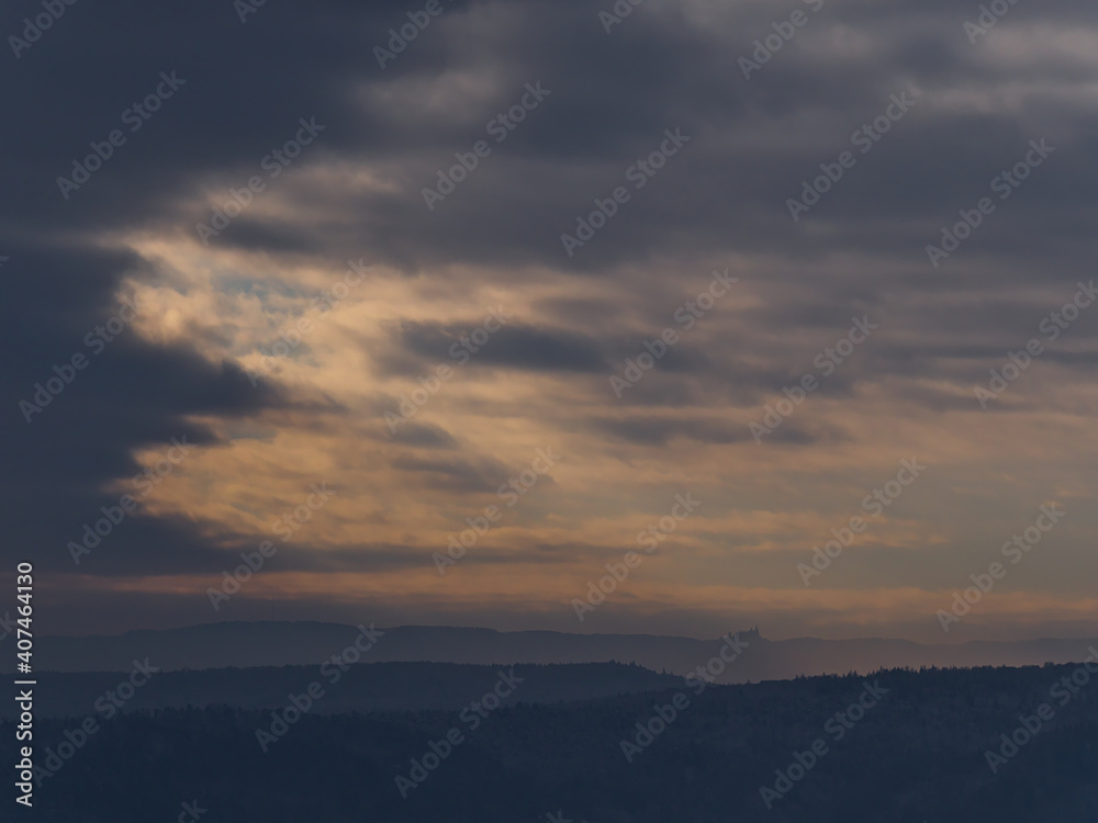 Silhouette of popular historic Hohenzollern Castle on the foothills of low mountain range Swabian Alb below dramatic brightening sky of orange colored clouds viewed from Wurmlingen, Germany in winter.