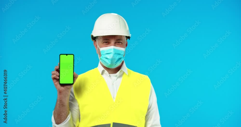 Portrait of happy middle-aged Caucasian man inspector in uniform and helmet wearing medical mask holding smartphone with green screen standing on blue wall in studio. Chroma key, builder concept Portrait of happy middle-aged Caucasian man inspector in uniform and helmet wearing medical mask holding smartphone with green screen standing on blue wall in studio. Chroma key, builder concept