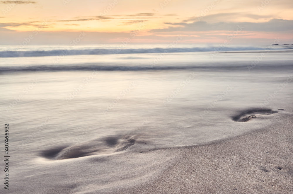 Footprints in the sand being washed away by a wave at sunset, long exposure photo, color toning applied, selective focus.