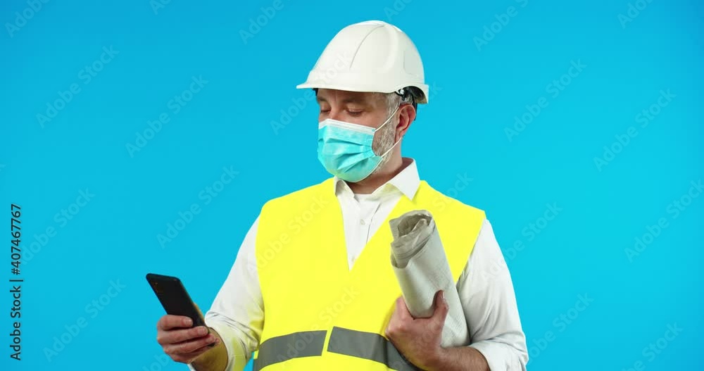 Portrait of serious Caucasian handsome middle-aged man professional builder in helmet in medical mask isolated on blue background with plan documents and typing on cellphone. Browsing on smartphone Portrait of serious Caucasian handsome middle-aged man professional builder in helmet in medical mask isolated on blue background with plan documents and typing on cellphone. Browsing on smartphone