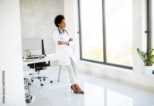 Female African American doctor wearing white coat with stethoscope standing by desk in office