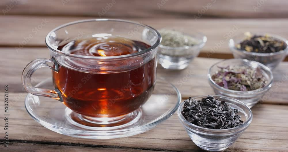 Drop slowly falls into a glass cup of tea. Around there are small bowls with different types of leaf tea. Vintage wooden table. Drop slowly falls into a glass cup of tea. Around there are small bowls with different types of leaf tea. Vintage wooden table.