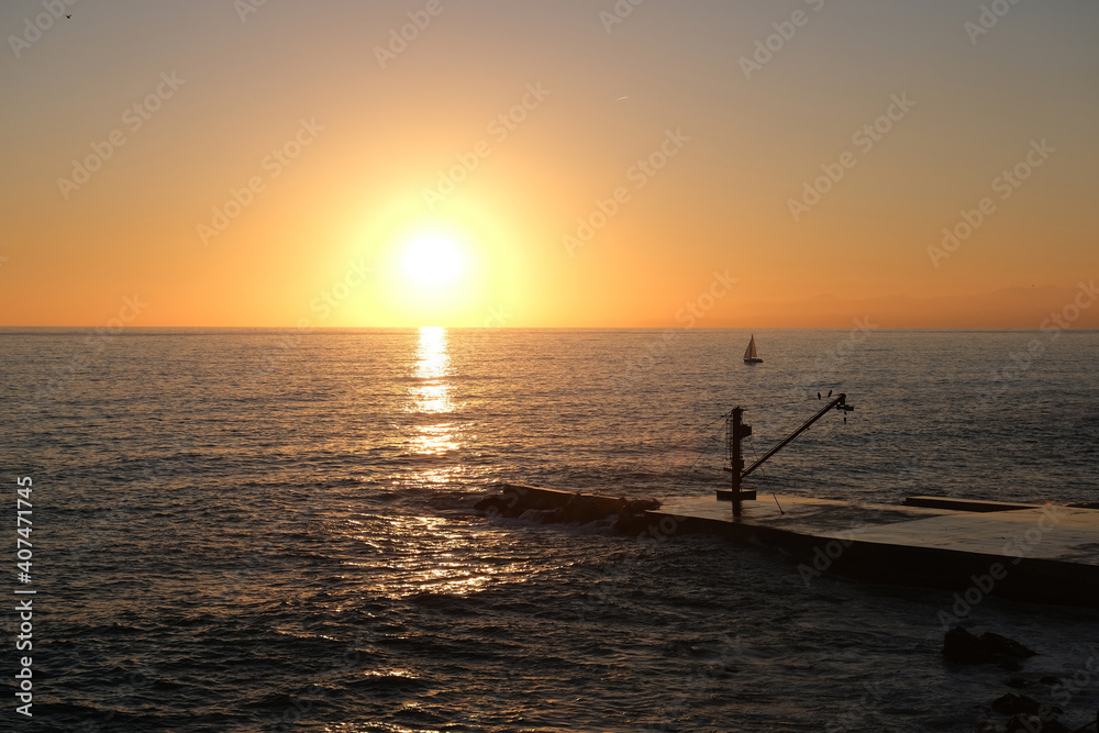 Un bellissimo tramonto sul mare a Genova con un piccolo molo ed una gru con un paio di cormorani appollaiati sopra ed una barca a vela in lontananza con il sole a pelo d'acqua che si tuffa nel mare
