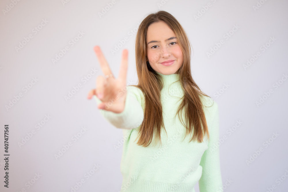 Young beautiful woman wearing green sweater standing over white isolated background smiling looking to the camera showing fingers doing victory sign. Number two. Young beautiful woman wearing green sweater standing over white isolated background smiling looking to the camera showing fingers doing victory sign. Number two.