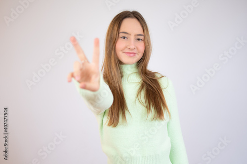 Young beautiful woman wearing green sweater standing over white isolated background smiling looking to the camera showing fingers doing victory sign. Number two.