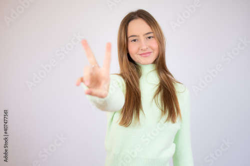 Young beautiful woman wearing green sweater standing over white isolated background smiling looking to the camera showing fingers doing victory sign. Number two.