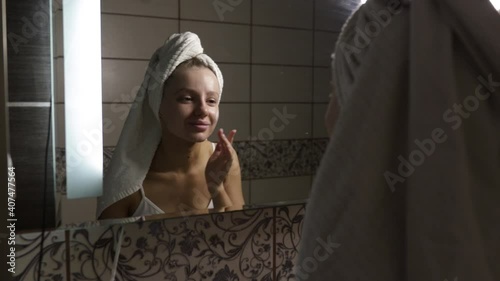 Beautiful Caucasian woman with a towel on her head in the bathroom applies cream after a shower to her face. 