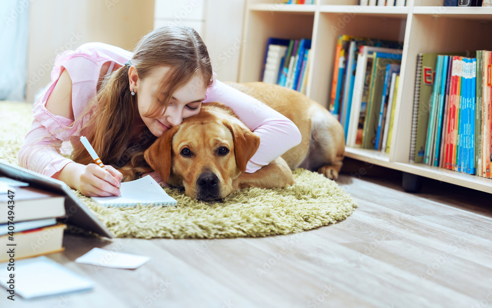 Teenage girl embracing pet dog while doing homework using tablet lying on carpet floor