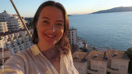 Cheerful solo female traveler filming a selfie video from her hotel balcony at the beach. Influencer concept.