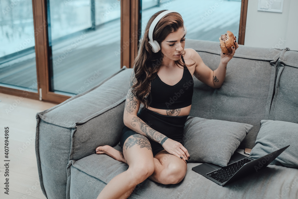 Obraz premium Beautiful sportswoman in black clothing with headphones and hamburger looks at laptop and sits on sofa in apartment.