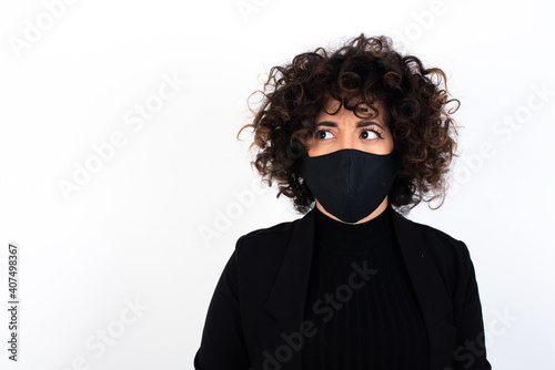 Amazed puzzled young beautiful caucasian woman wearing medical mask standing against white wall, curves lips and has worried look, sees something awful in front.