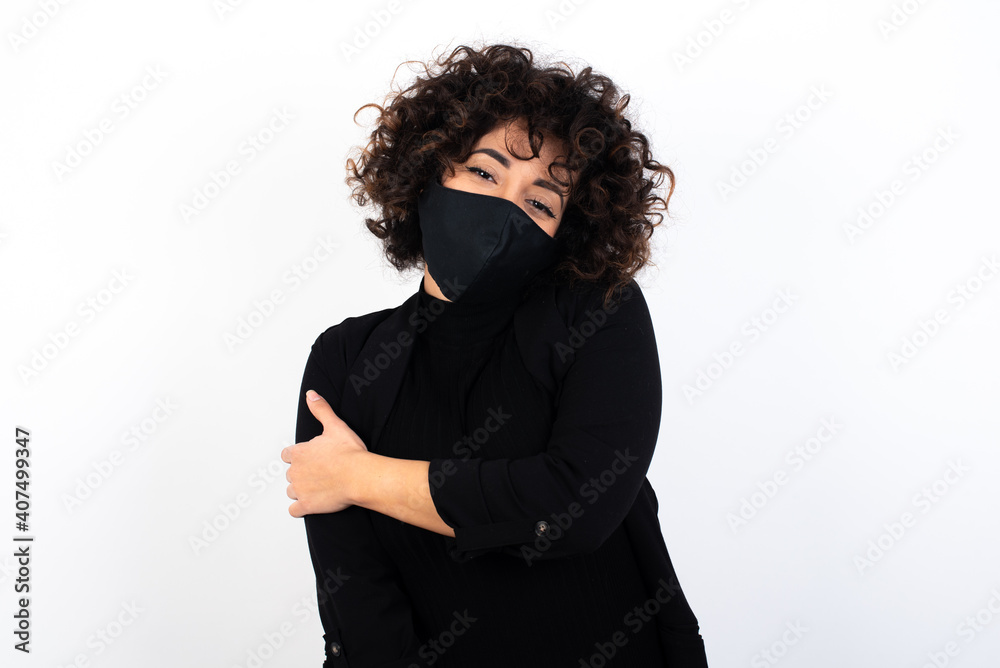 Fototapeta premium People, lifestyle, youth and happiness concept. Shy pretty young beautiful caucasian woman wearing medical mask standing against white wall,, feeling happy hugging herself.