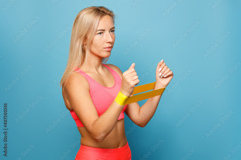 Portrait of slim fitness girl stands and stretches an elastic band with her hands on a blue background. Portrait of slim fitness girl stands and stretches an elastic band with her hands on a blue background.