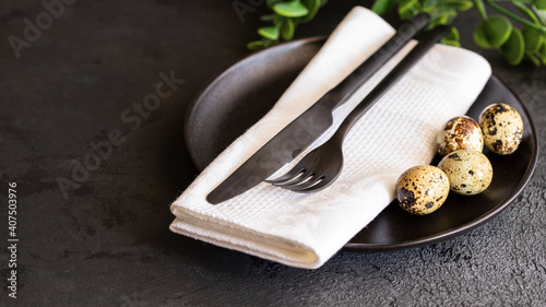 Happy Easter concept. Black ceramic plate with cutlery, quail eggs, white napkin and green plant for festive dinner. Modern table setting  for celebration photo