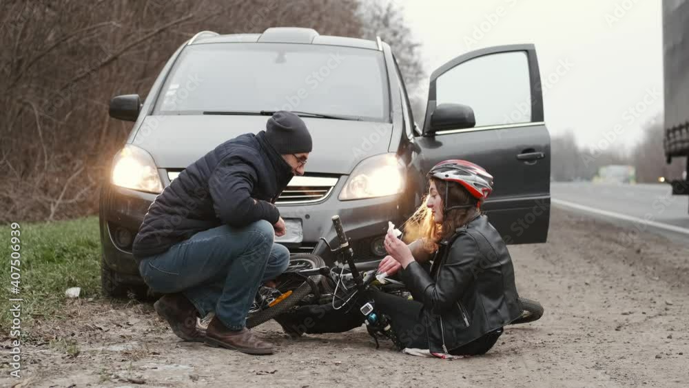 Female cyclist injured on the street bike after collision accident car and bike. Woman with nose bleeding sitting on the road and man giving her first aid, medicaments.