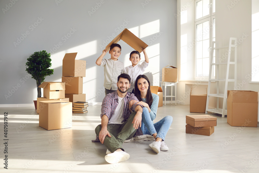 Portrait of happy young family in new home. Smiling mom and dad sitting on floor with unpacked boxes, and children holding symbolic roof behind. Real estate, mortgage, buying and moving house concept Portrait of happy young family in new home. Smiling mom and dad sitting on floor with unpacked boxes, and children holding symbolic roof behind. Real estate, mortgage, buying and moving house concept