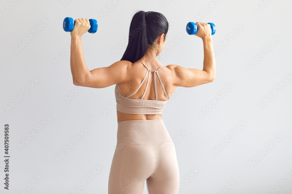 Back view of female model posing in studio and demonstrating comfortable activewear. Rear view of young woman in modern light beige close-fitting sportswear holding blue dumbbells and showing fit body Back view of female model posing in studio and demonstrating comfortable activewear. Rear view of young woman in modern light beige close-fitting sportswear holding blue dumbbells and showing fit body