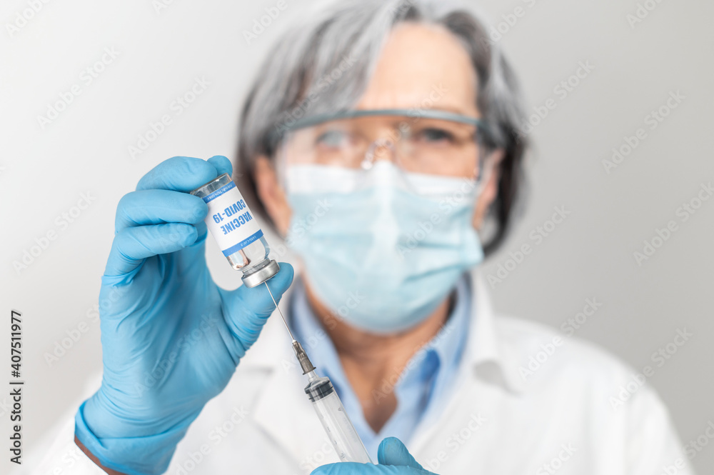 Close-up image of covid-19 vaccine, blurred senior female doctor, healthcare professional wearing protective face mask, goggles and gloves holding and showing created vaccine from coronavirus Close-up image of covid-19 vaccine, blurred senior female doctor, healthcare professional wearing protective face mask, goggles and gloves holding and showing created vaccine from coronavirus