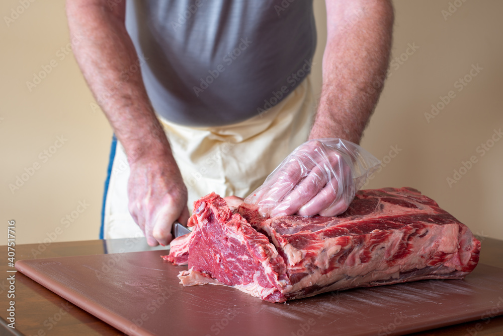 A male butcher or chef is preparing prime rib steaks from a large prime beef rib roast with multiple bones. The meat is on a brown plastic cutting board which is sitting on a restaurant counter. 