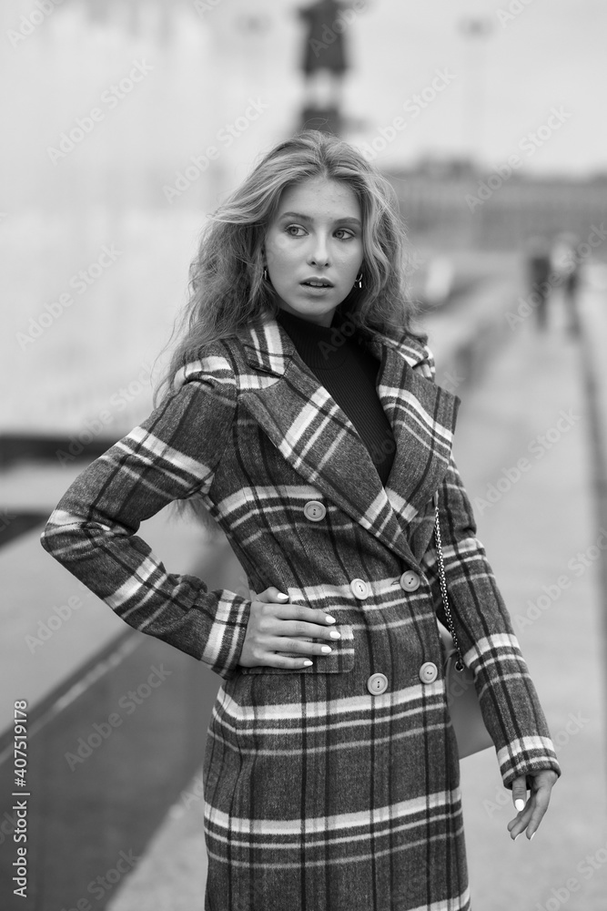 Fototapeta premium Elegant woman walking city street. Fashion fall autumn look. Pretty caucasian blonde stylish girl wearing blue jeans, wine red pullover and squared wool coat. Female with makeup and wavy blonde hair
