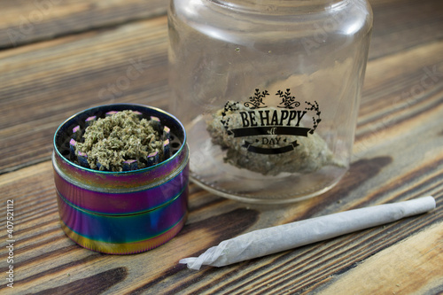 „Be happy all day” jar with marijuana buds and joint lie on a wooden background. Grinder near cannabis. Marijuana is medicine.