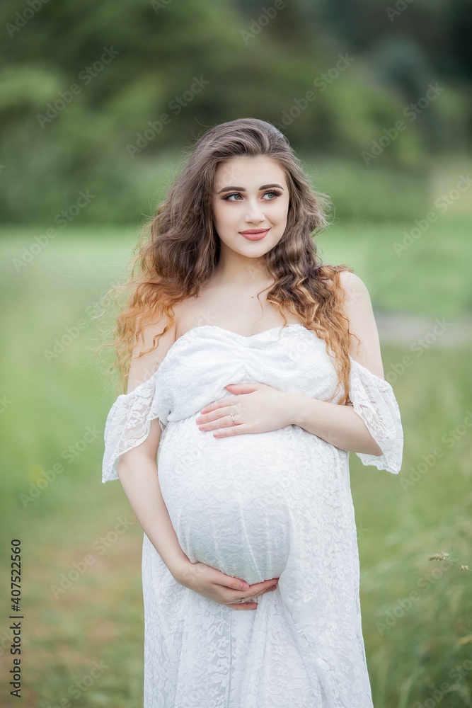 A beautiful young pregnant woman with curly long hair in a white dress walks in the field. Spring portrait of a pregnant woman. Happy pregnancy. A beautiful young pregnant woman with curly long hair in a white dress walks in the field. Spring portrait of a pregnant woman. Happy pregnancy.