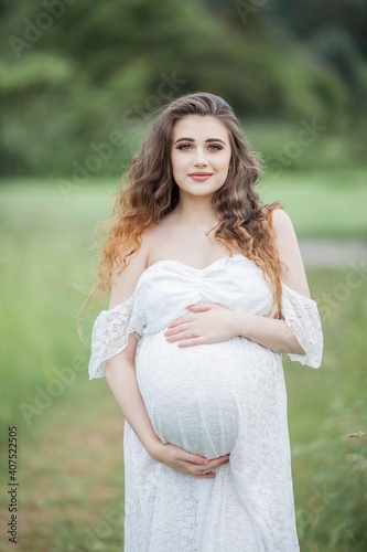 A beautiful young pregnant woman with curly long hair in a white dress walks in the field. Spring portrait of a pregnant woman. Happy pregnancy.