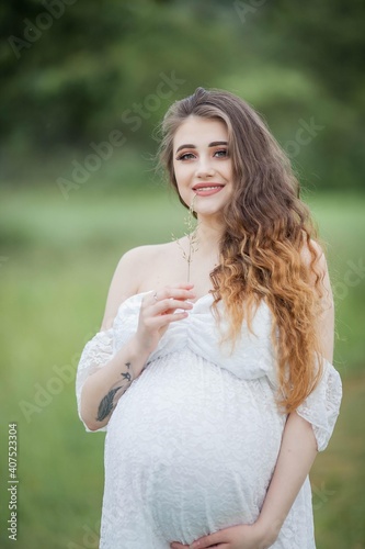 Fototapeta Naklejka Na Ścianę i Meble -  A beautiful young pregnant woman with curly long hair in a white dress walks in the field. Spring portrait of a pregnant woman. Happy pregnancy.