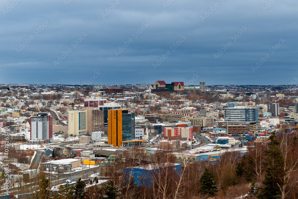 A cityscape of the City of St. John's, Newfoundland on a cloudy day. There are a number of large multi-story buildings both residential and commercial. The trees are bare and snow is on the ground.