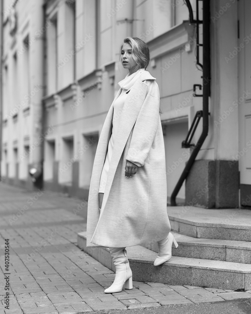 Elegant woman in white dress, hessian boots and coat walking at city street. Fall autumn fashion look. Pretty tall stylish young girl with fashionable makeup and hair style. Full length portrait