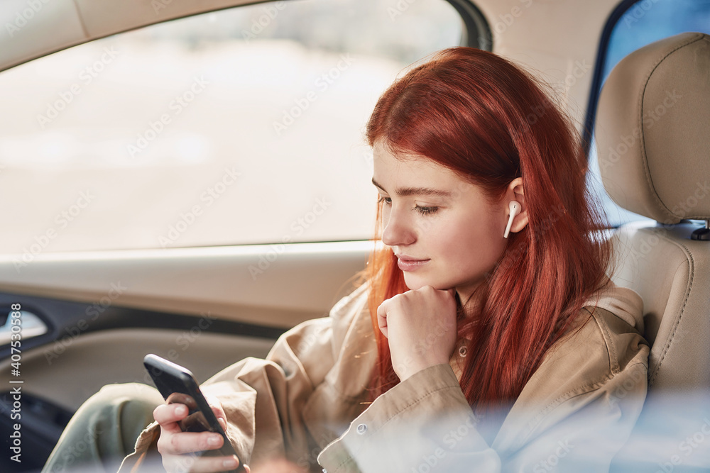 Teenage girl in earphones looking at the screen with a smile, while using her phone, watching videos sitting in the car Teenage girl in earphones looking at the screen with a smile, while using her phone, watching videos sitting in the car