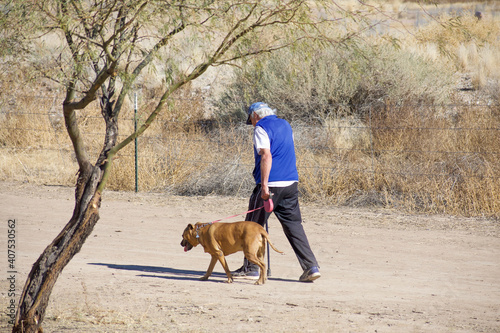 Elderly masked man walking his dog along desert trail during pandemic.