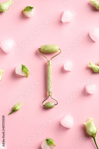 Green jade stone face roller top view on pink background with ice cubes and flowers.
