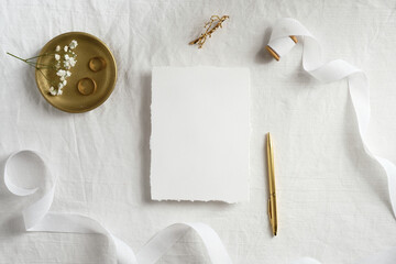 Blank paper card mockup, white ribbon, golden pen and rings on textile background. Top view, flat lay. Wedding invitation template.