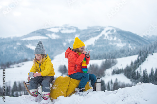Winter children travellers enjoy coffee time with scenery view of the snowy mountain background. Kids in snow drink tea.