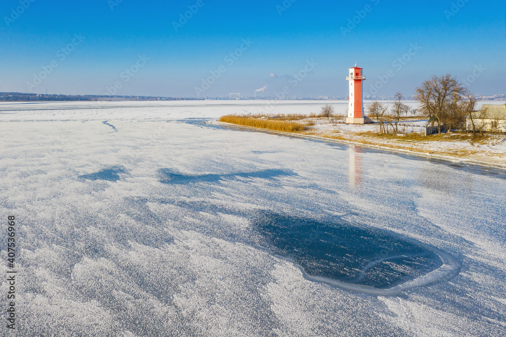 aerial view to frozen river with ice and alone red lighthouse on the coast under blue sky in winter day
