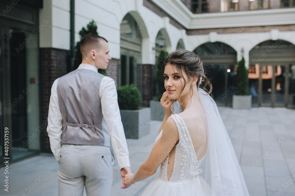 The brunette curly-haired bride turned around in a white dress with a long veil smiling, looking into the frame, walking in the city, holding the groom's hand. Wedding portrait of the newlyweds.