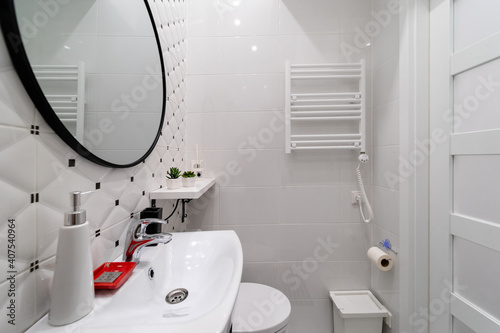 A part of a modern small bathroom in a newly furnished studio apartment in an old house. Ceramic tiled walls, towel radiator on white wall, sink, mirror, shelf with flowers, door, toilet, laundry bag